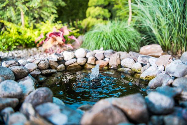 An elegant small garden fountain surrounded by vibrant greenery. The fountain features a tiered design with water cascading gently down, creating a tranquil atmosphere. Colorful flowers and lush plants frame the fountain, adding to the serene garden setting.